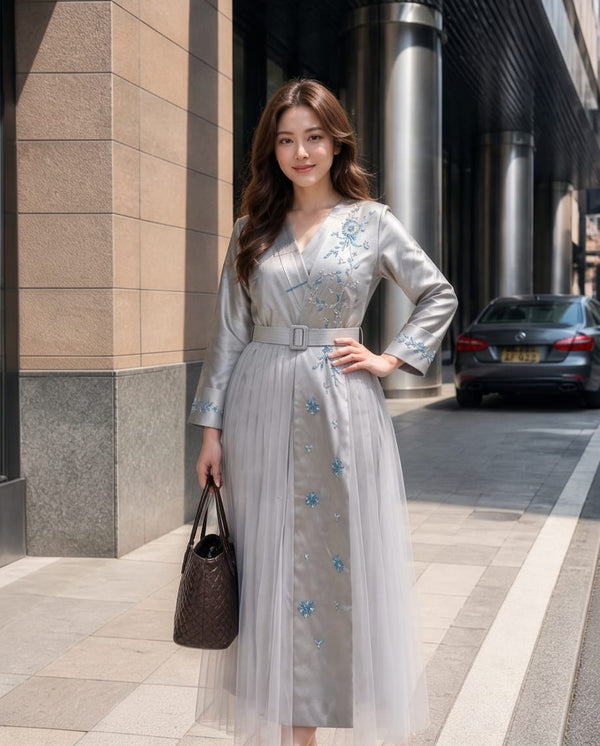 A woman stands on a city sidewalk wearing a long, light gray tulle dress with blue floral embroidery and a matching belt. She is holding a dark brown woven handbag, with a modern building facade and a parked car in the background.